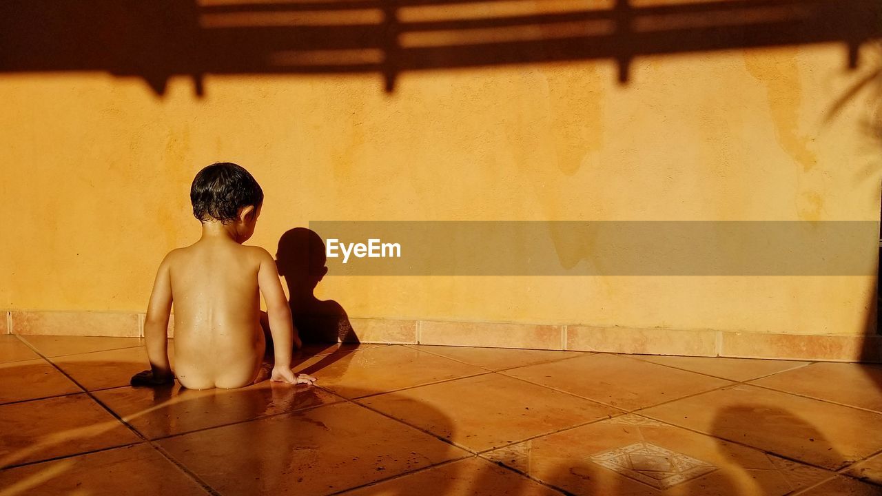Rear view of naked boy sitting on floor at home