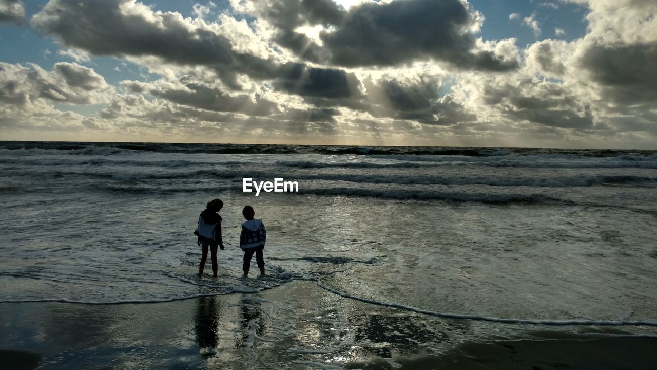 Rear view of friends standing on shore at beach against cloudy sky