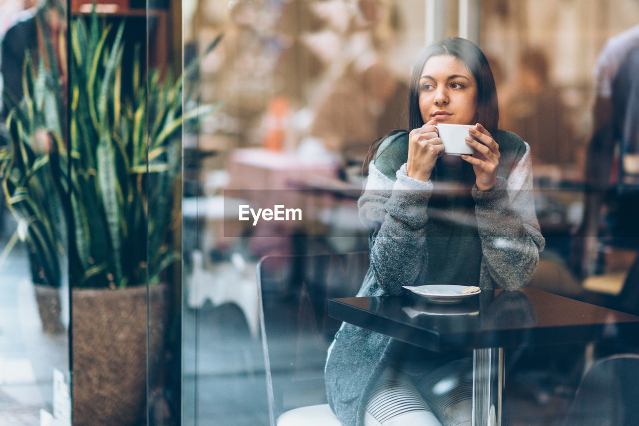Thoughtful young woman holding coffee cup while looking through window in cafe