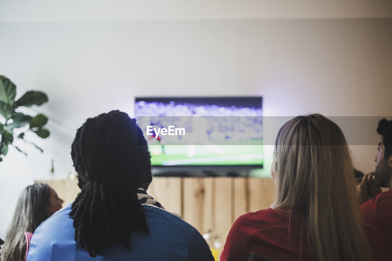 Rear view of friends watching soccer match while sitting at living room