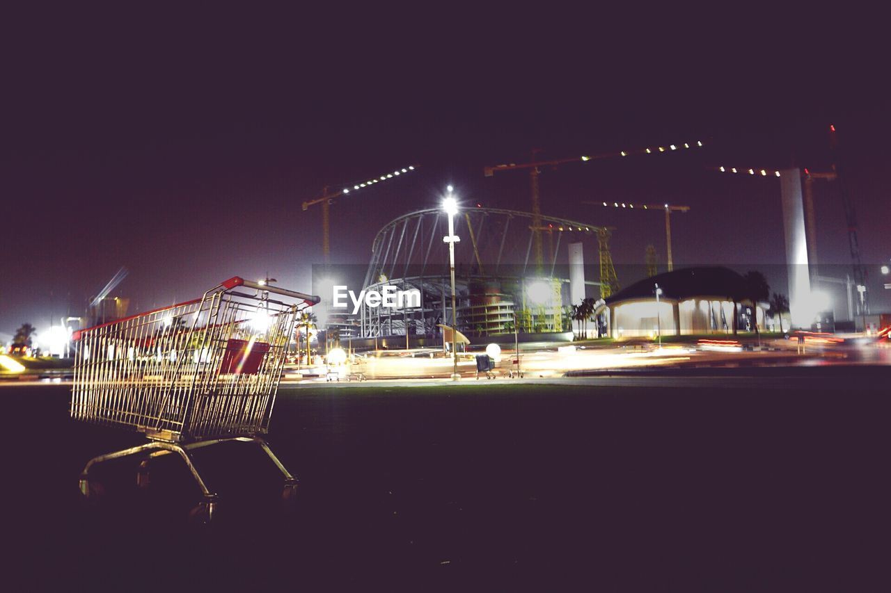 Shopping cart on field against illuminated construction site at night