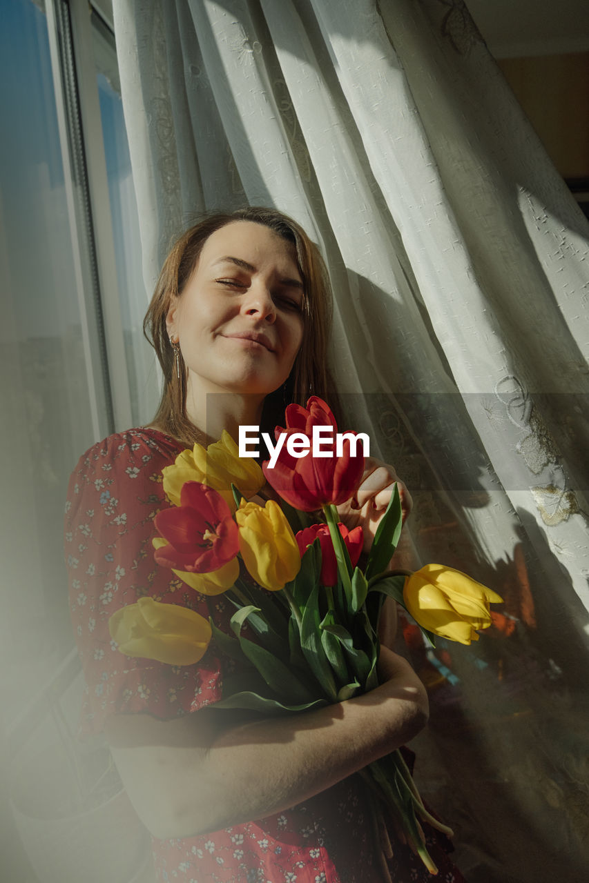 Natural beauty young woman in red dress with blooming tulips in room interior near windows, sunlight 