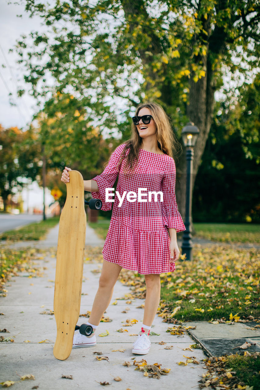 Girl smiling wearing sunglasses standing on sidewalk holding a skateboard