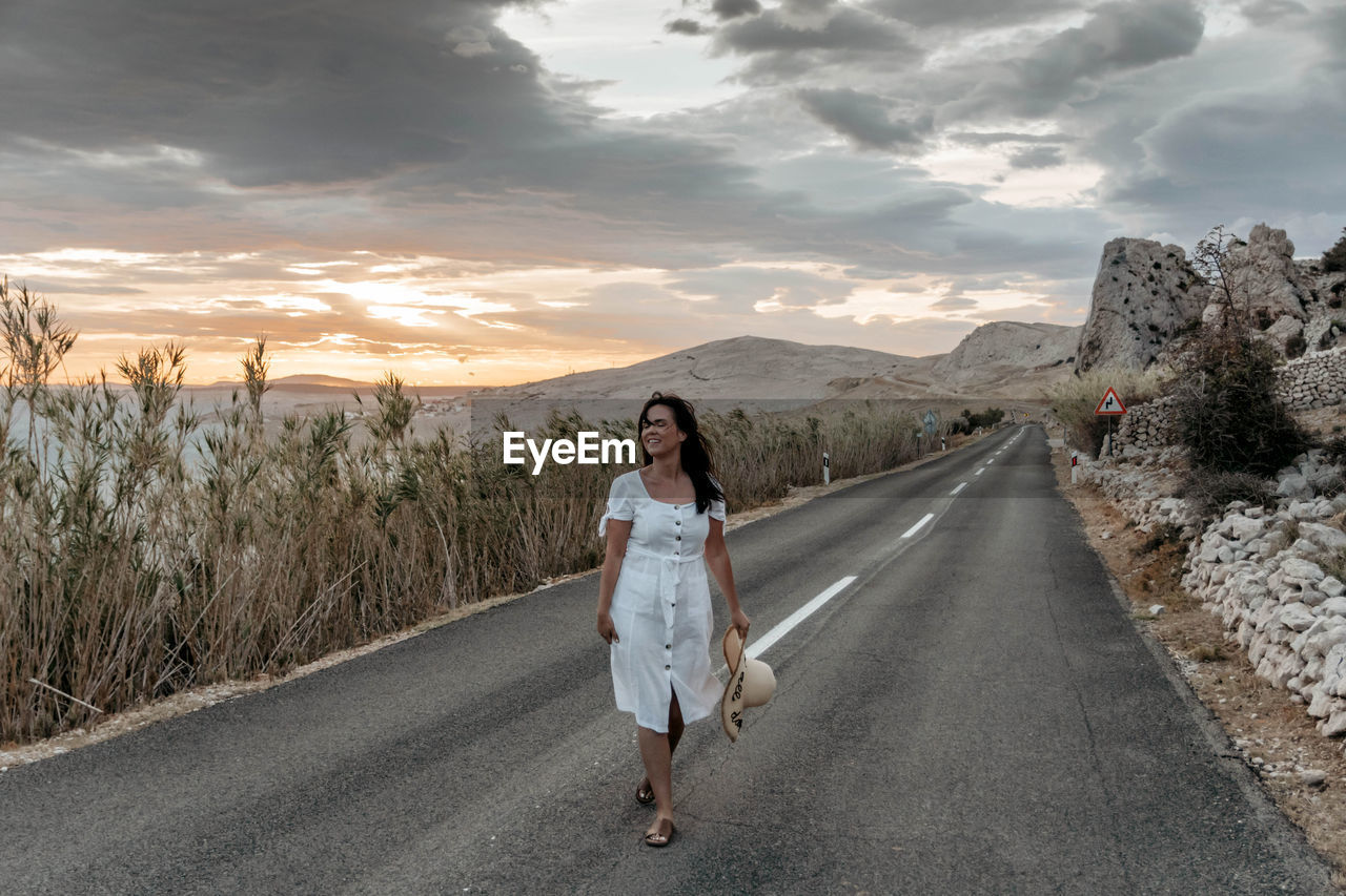 Front view of beautiful young woman in white dress standing on open road.