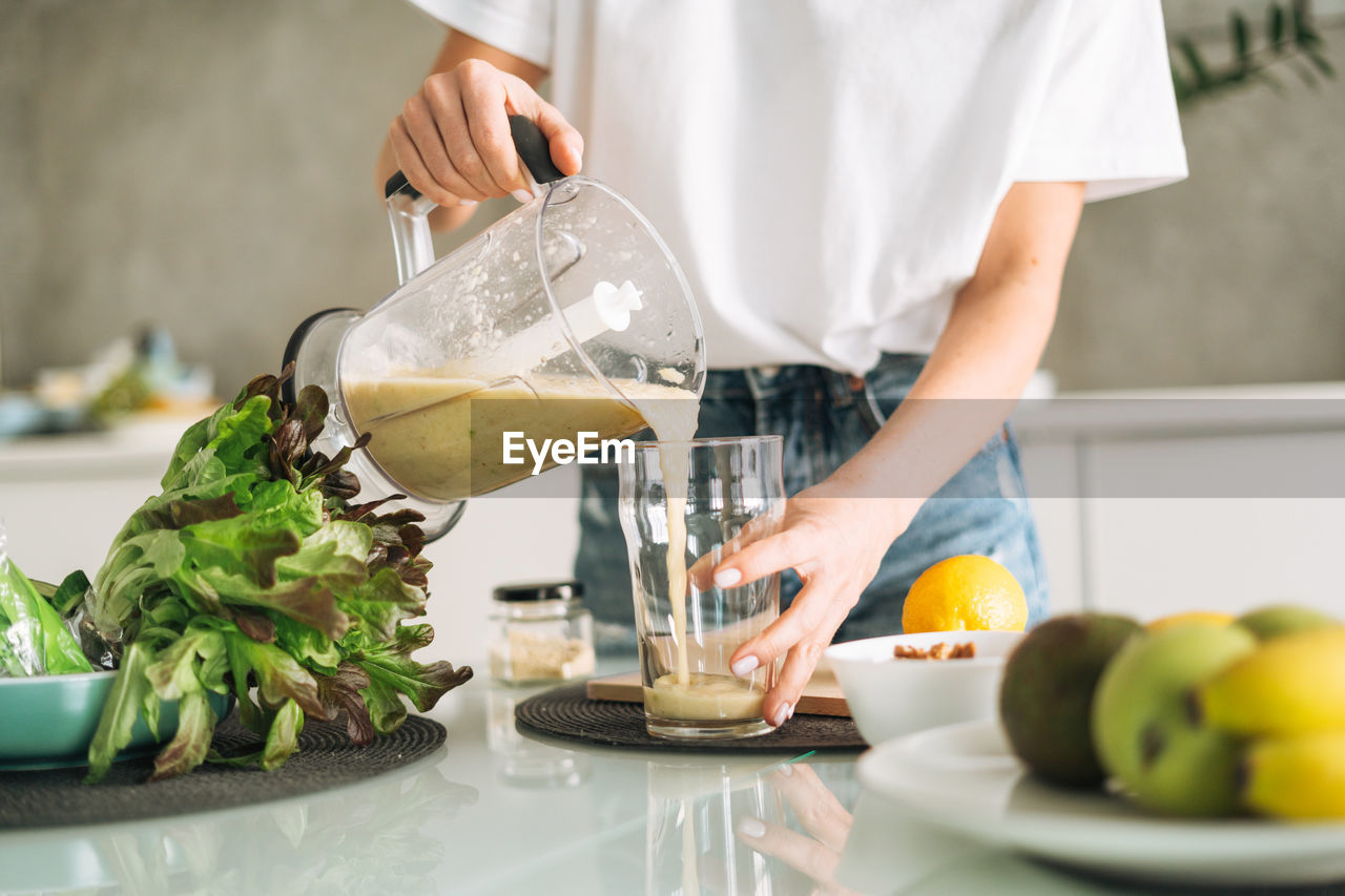 Young slim woman in white t-shirt and blue jeans cooking green smoothie healthy food in kitchen 