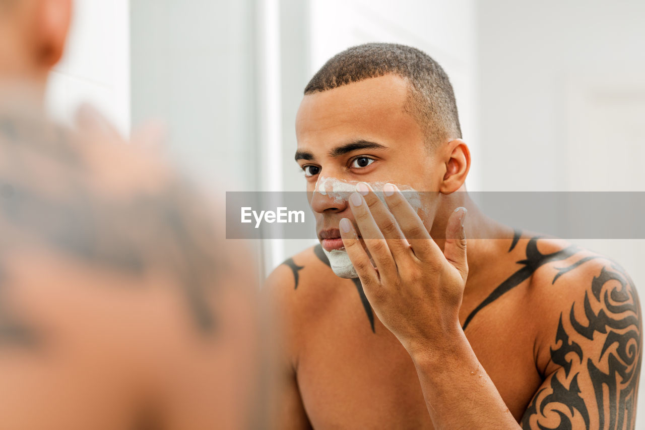 Tattooed shirtless man applying shaving cream on face while looking in mirror