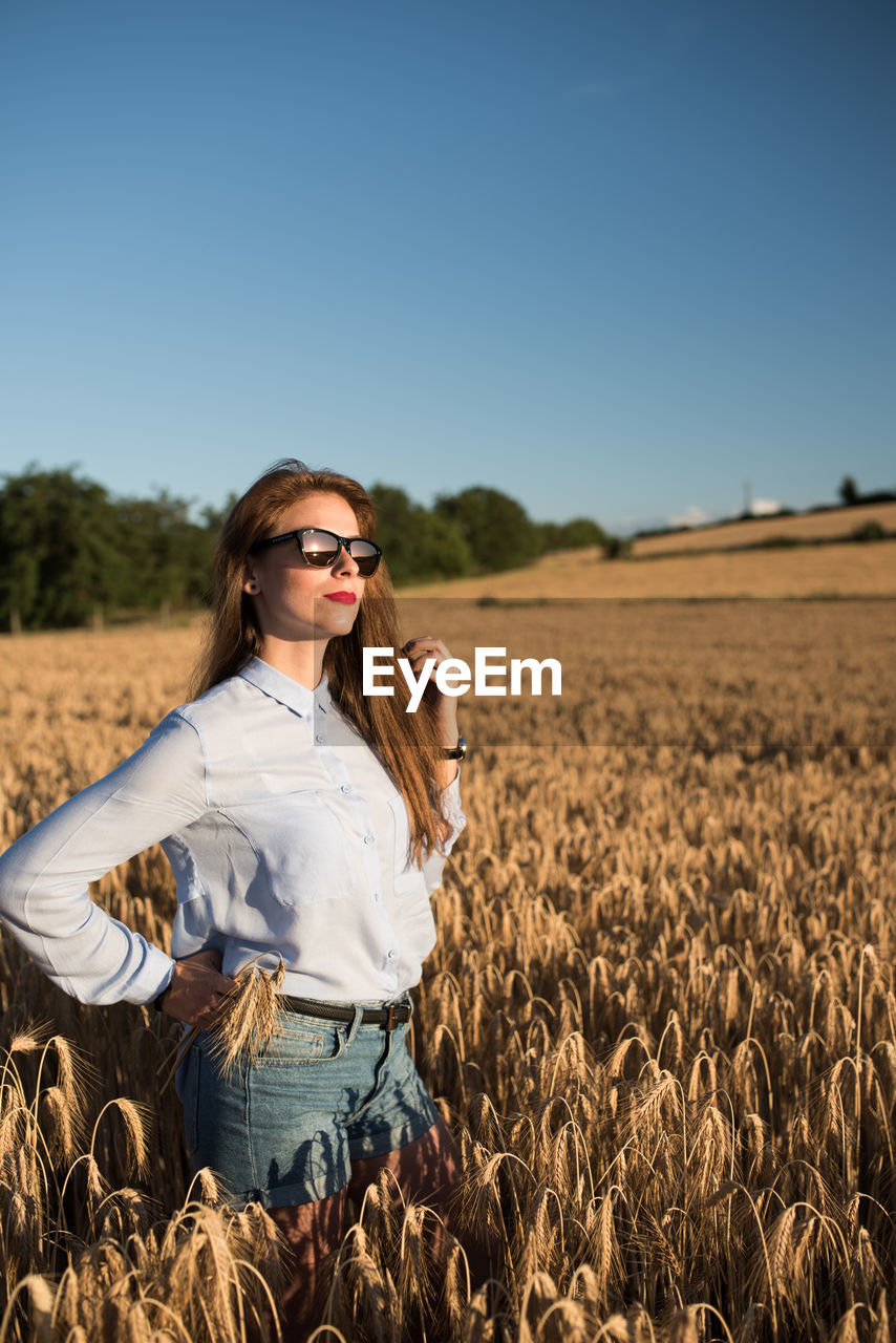 Beautiful young woman standing on wheat field against clear sky