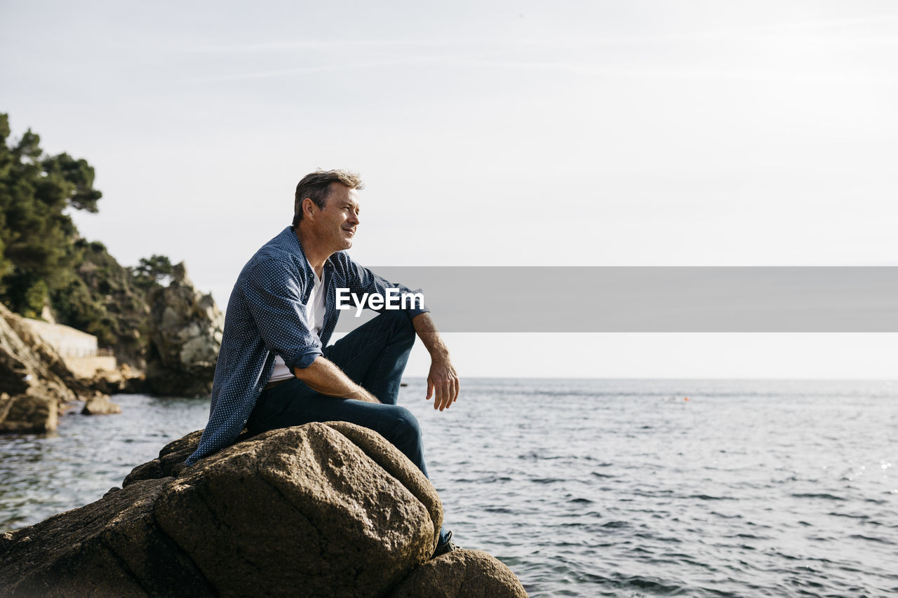 Thoughtful mature man sitting on rock against clear sky