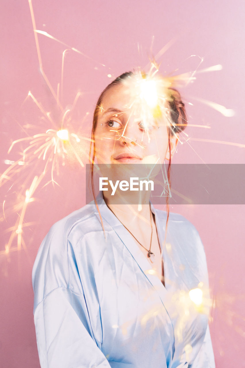 Digital composite image of thoughtful young woman and sparks against pink background