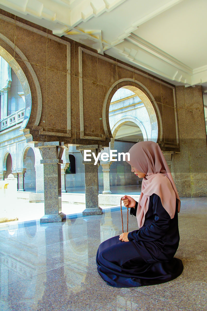 Full length of woman in hijab holding counting rosary beads while praying at mosque