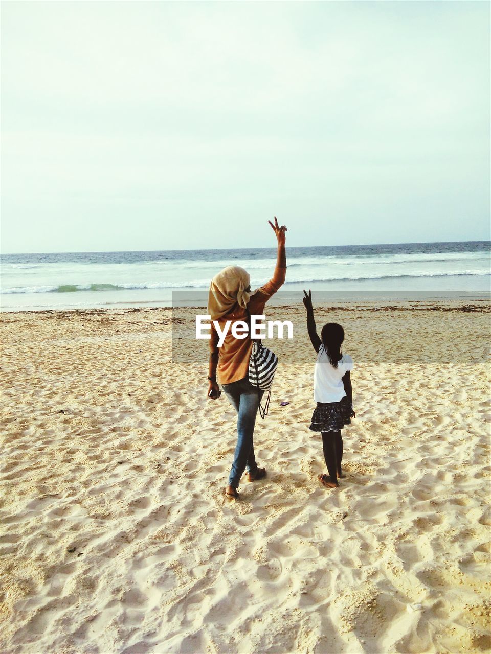 Rear view of woman and girl gesturing peace sign while standing on sand at beach against sky