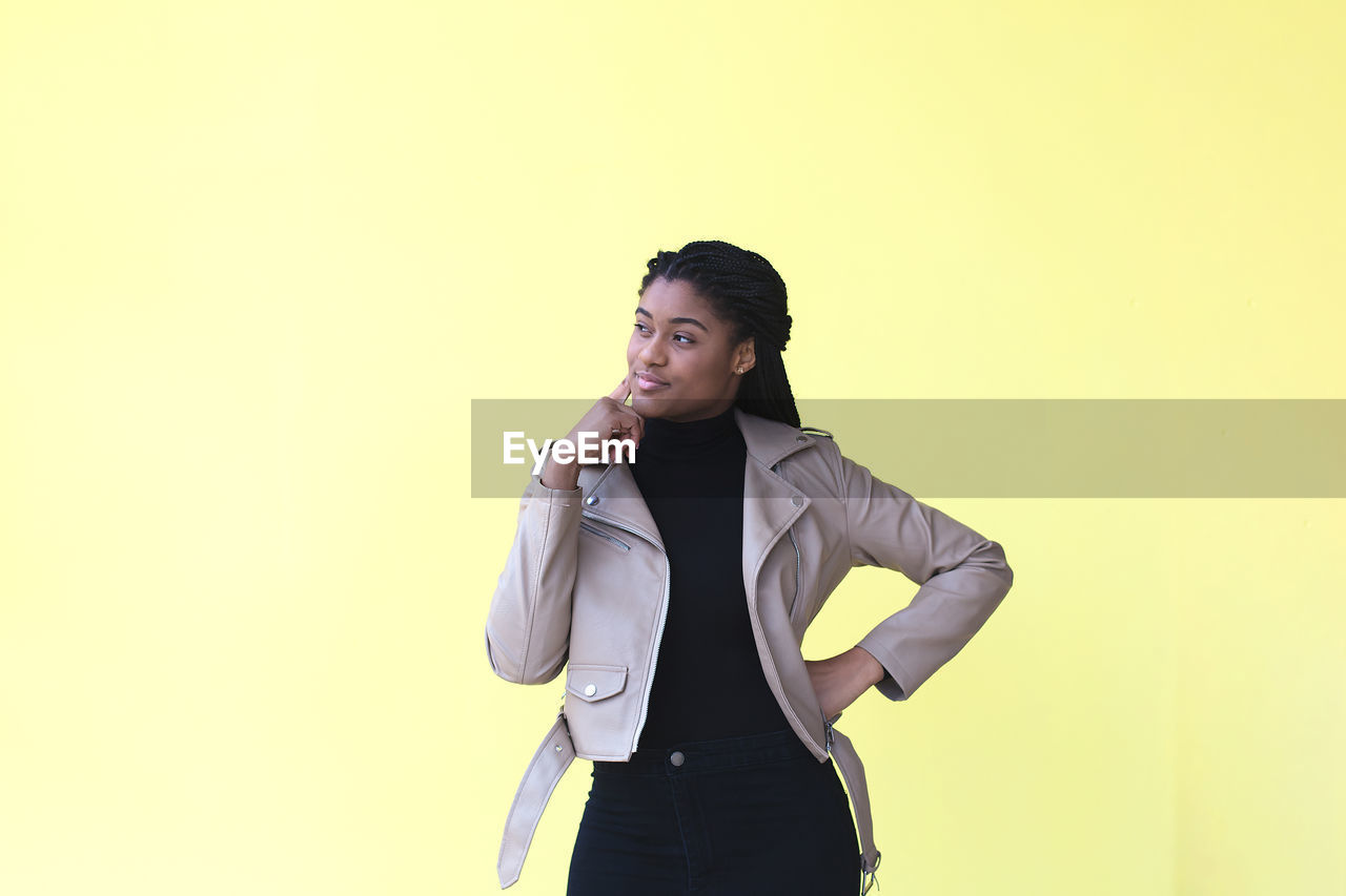 YOUNG WOMAN LOOKING AWAY WHILE STANDING AGAINST YELLOW WALL