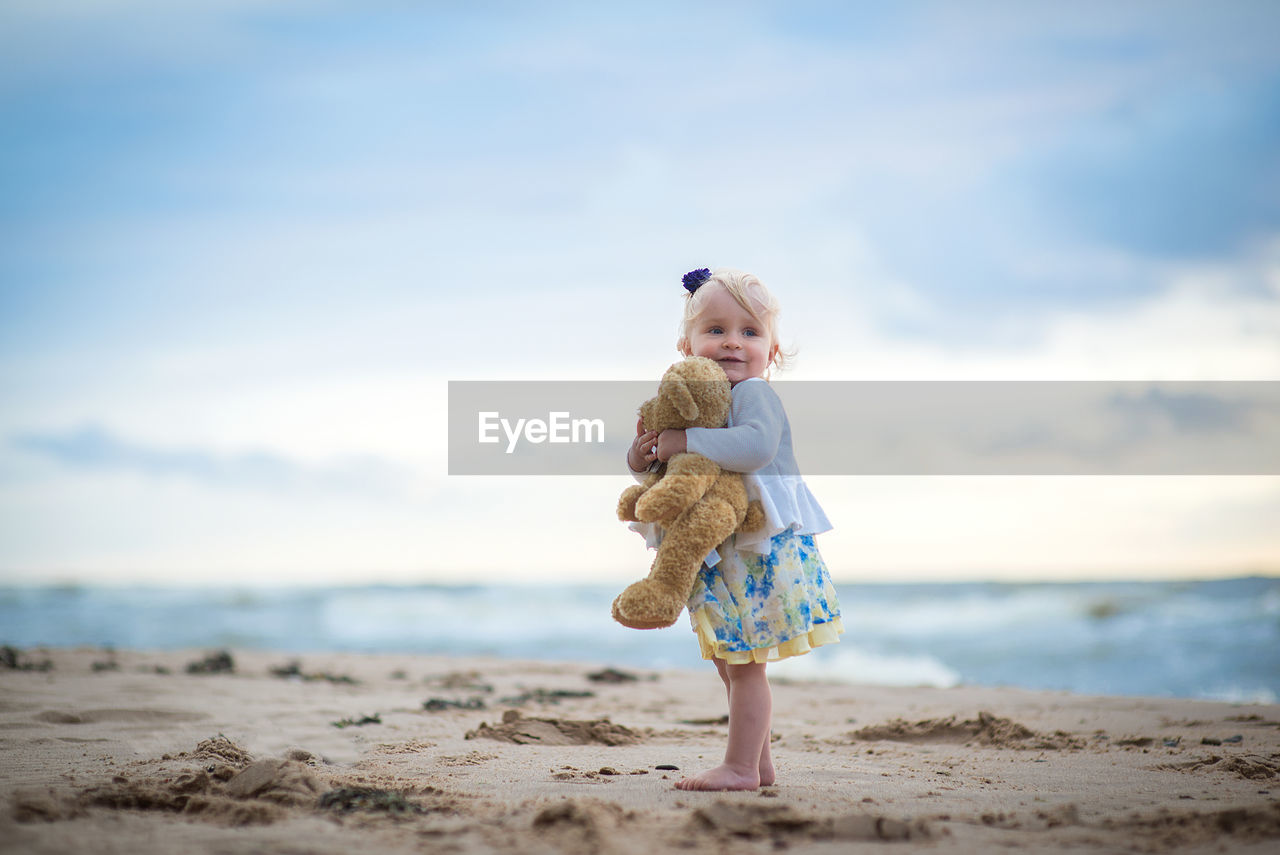 Cute girl holding stuffed toy while standing against sea at beach