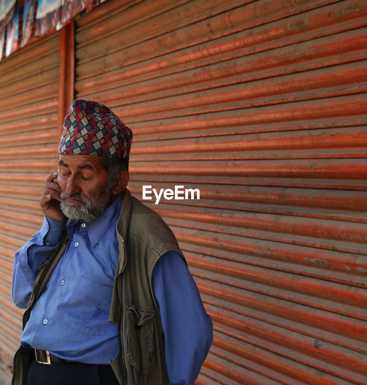 Senior man wearing dhaka topi listening to mobile phone against shutters