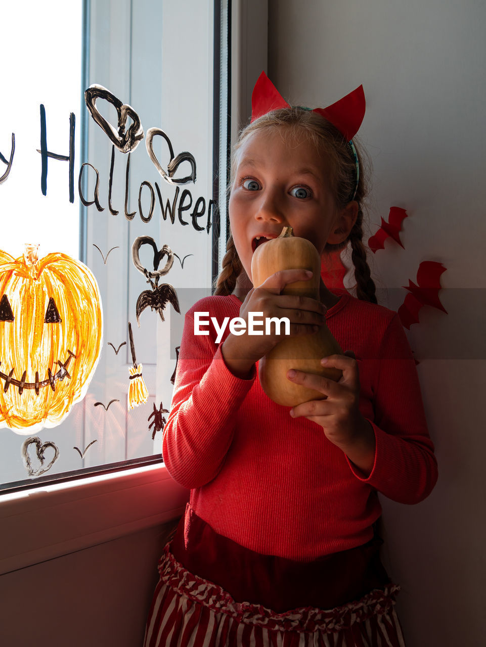 Child chewing pumpkin painting on window preparing to celebrate halloween little girl decorates room
