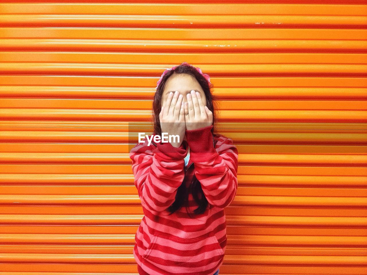 Front view of girl covering face with hands while standing against corrugated iron