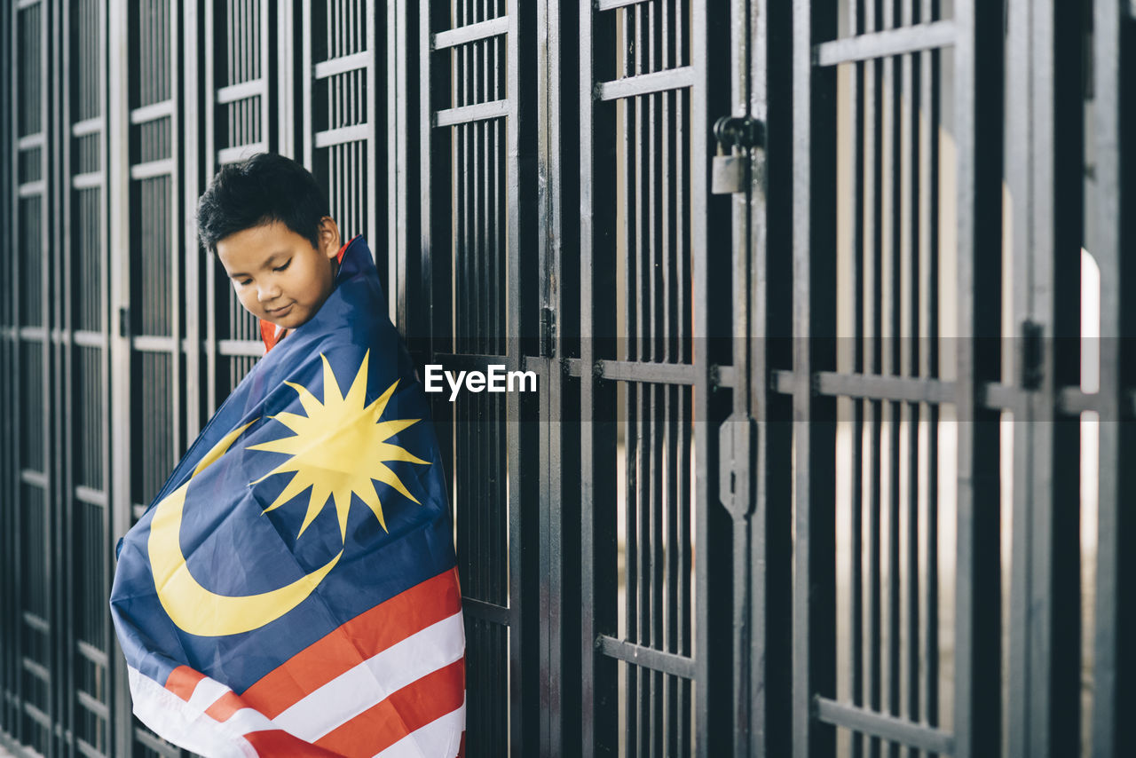 Side view of boy wearing malaysian flag by fence