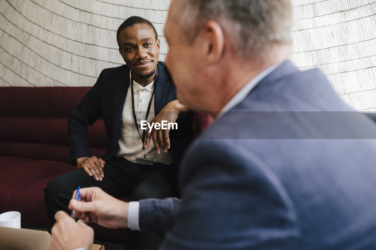 Smiling businessman looking at male colleague discussing while sitting on sofa during networking event