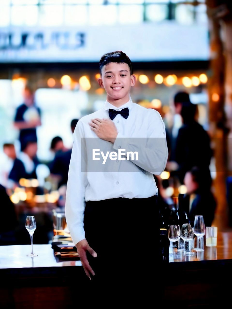 adult, food and drink, waiter, business, bar, refreshment, smiling, drink, service, restaurant, one person, men, glass, portrait, alcohol, standing, bar counter, happiness, young adult, bartender, food and drink industry, bow tie, looking at camera, emotion, nightlife, focus on foreground, happy hour, drinking glass, front view, cheerful, night, occupation, elegance, clothing, cafe, city, luxury, waist up, wine, table, three quarter length, formal wear, food, relaxation, fashion, holding, indoors, wealth, city life, wine glass, uniform