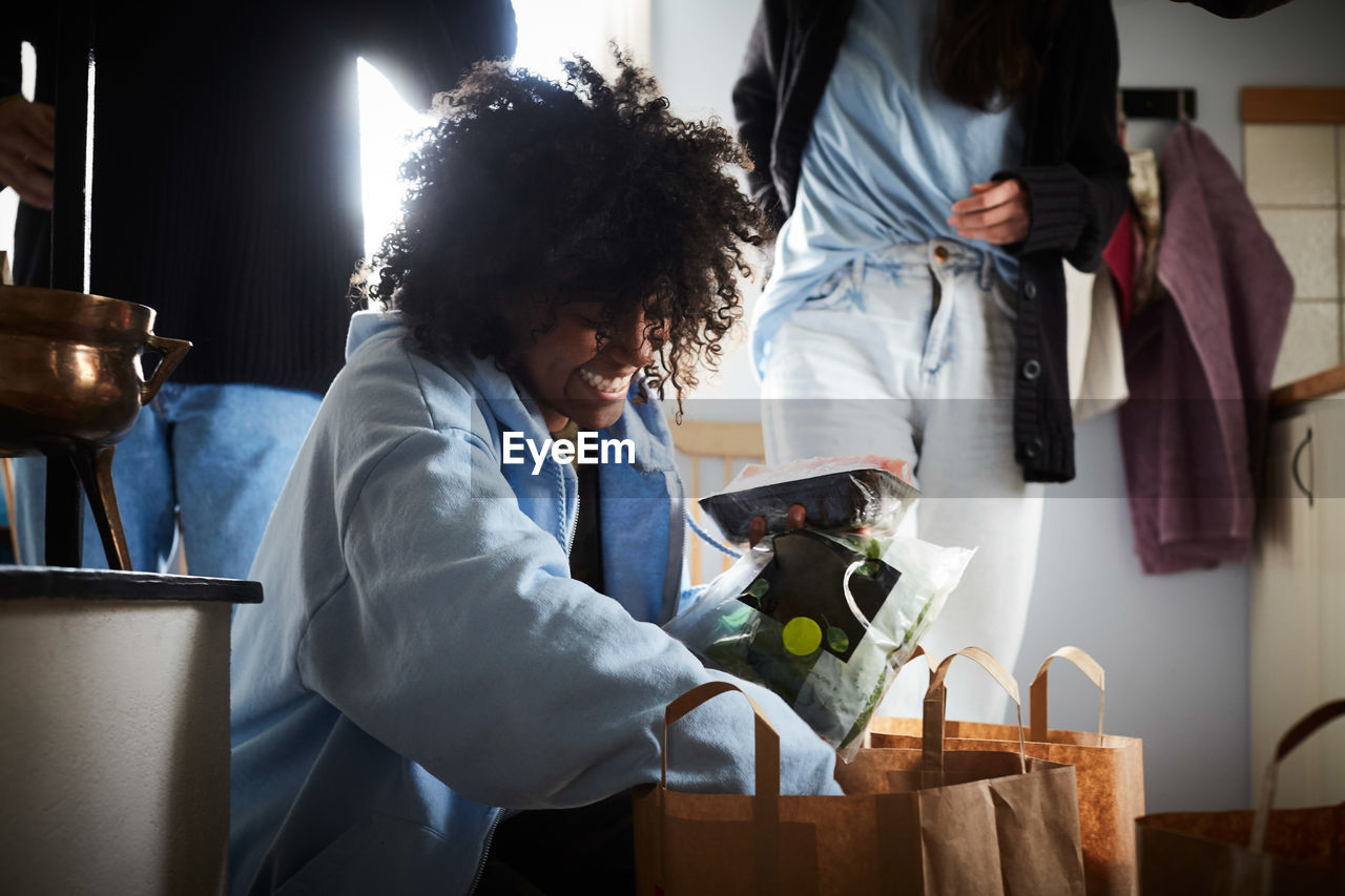 Smiling young woman removing groceries from shopping bags while friends standing in background at home