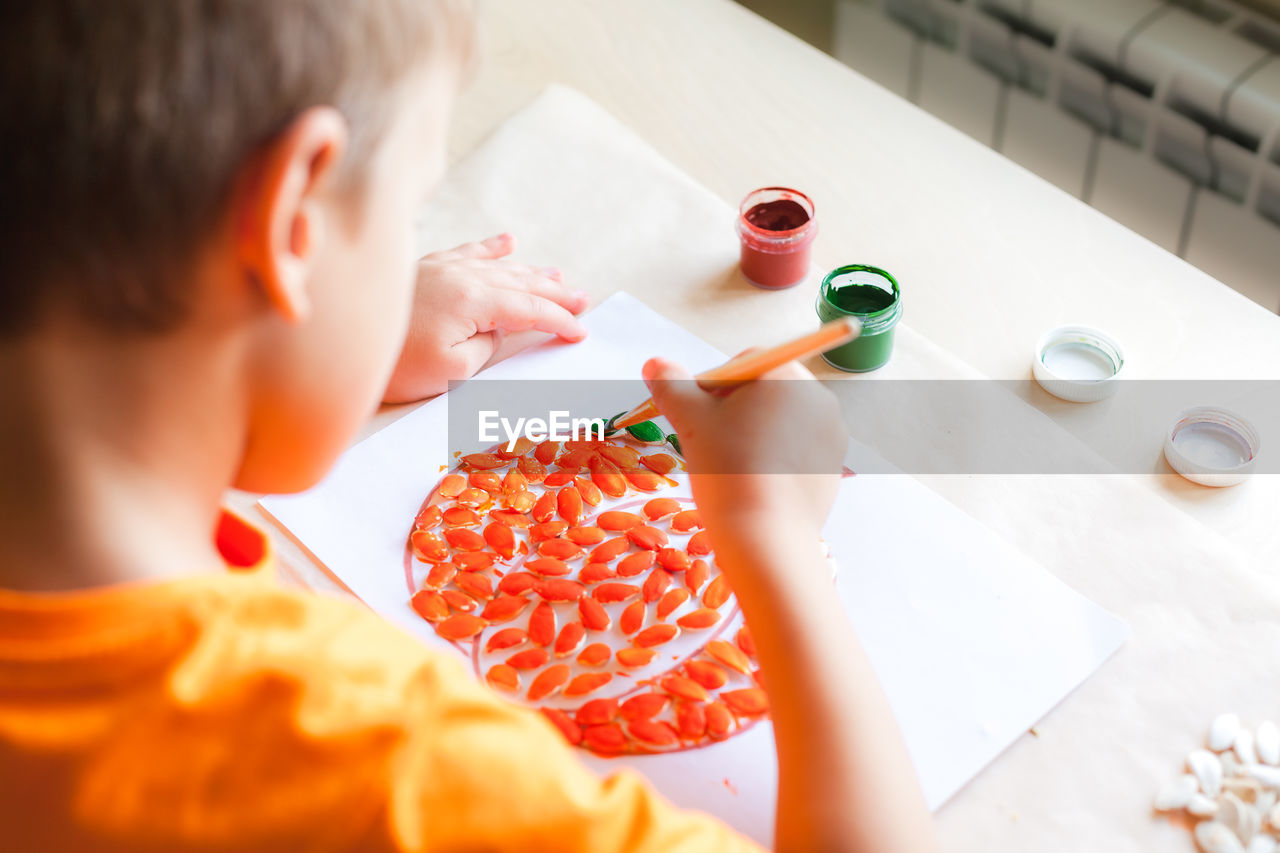 Cute child boy making pumpkin from seeds on white paper, halloween diy concept