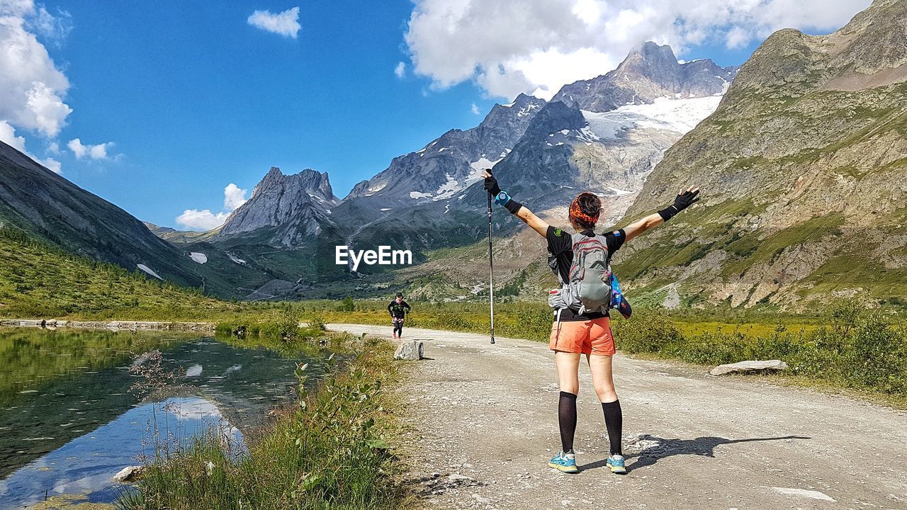 Rear view of mid adult woman with arms outstretched standing on road amidst mountains against sky