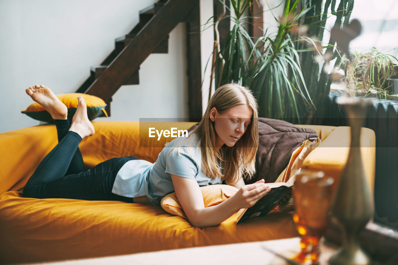 Woman reading book while lying on sofa at home