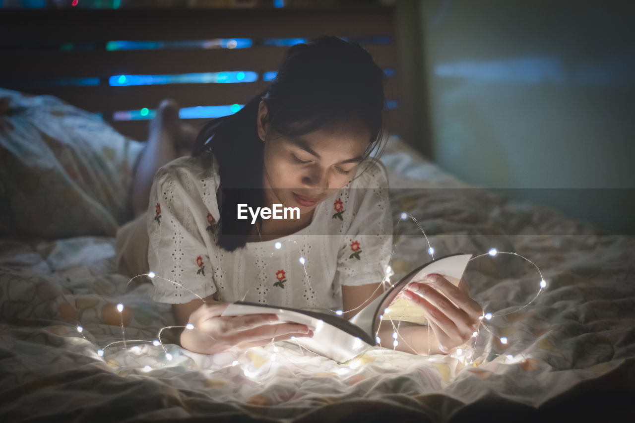 Young woman is reading a book with led lights on the bed in her bedroom.