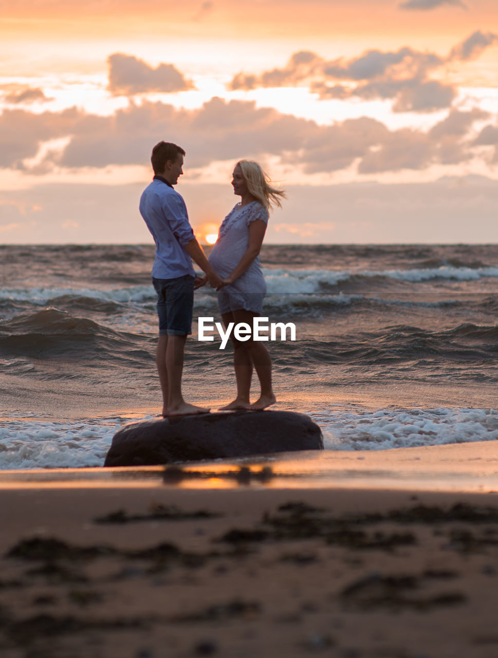 Side view of expectant couple standing on shore at beach during sunset