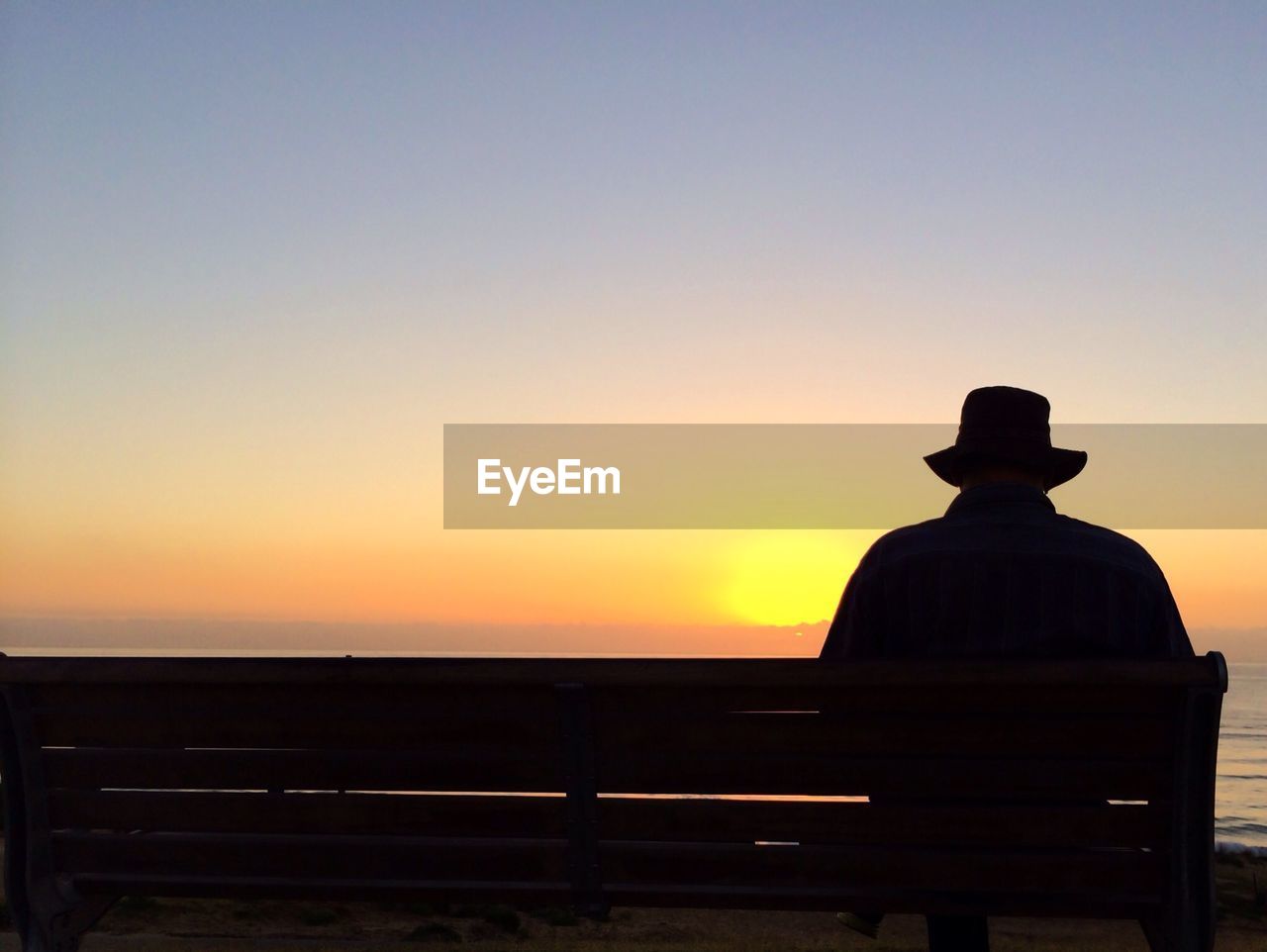 Rear view of silhouette man sitting on bench at sunset