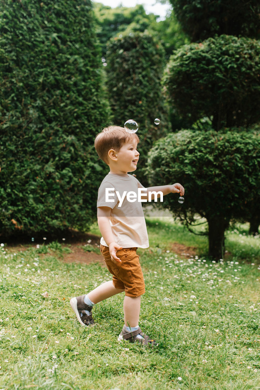 Cute happy little baby boy is running on the grass in the park trying to catch a soap bubble