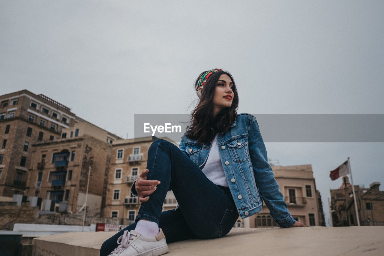 PORTRAIT OF BEAUTIFUL YOUNG WOMAN SITTING OUTDOORS AGAINST SKY