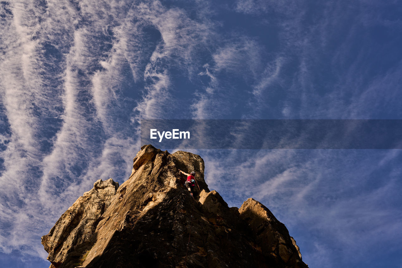 Low angle full length of active senior woman climbing on rocky cliff against sky