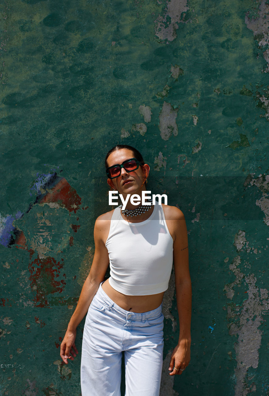 Confident young ethnic female model with dark hair on fashionable sunglasses and looking at camera leaning on shabby wall on sunny day