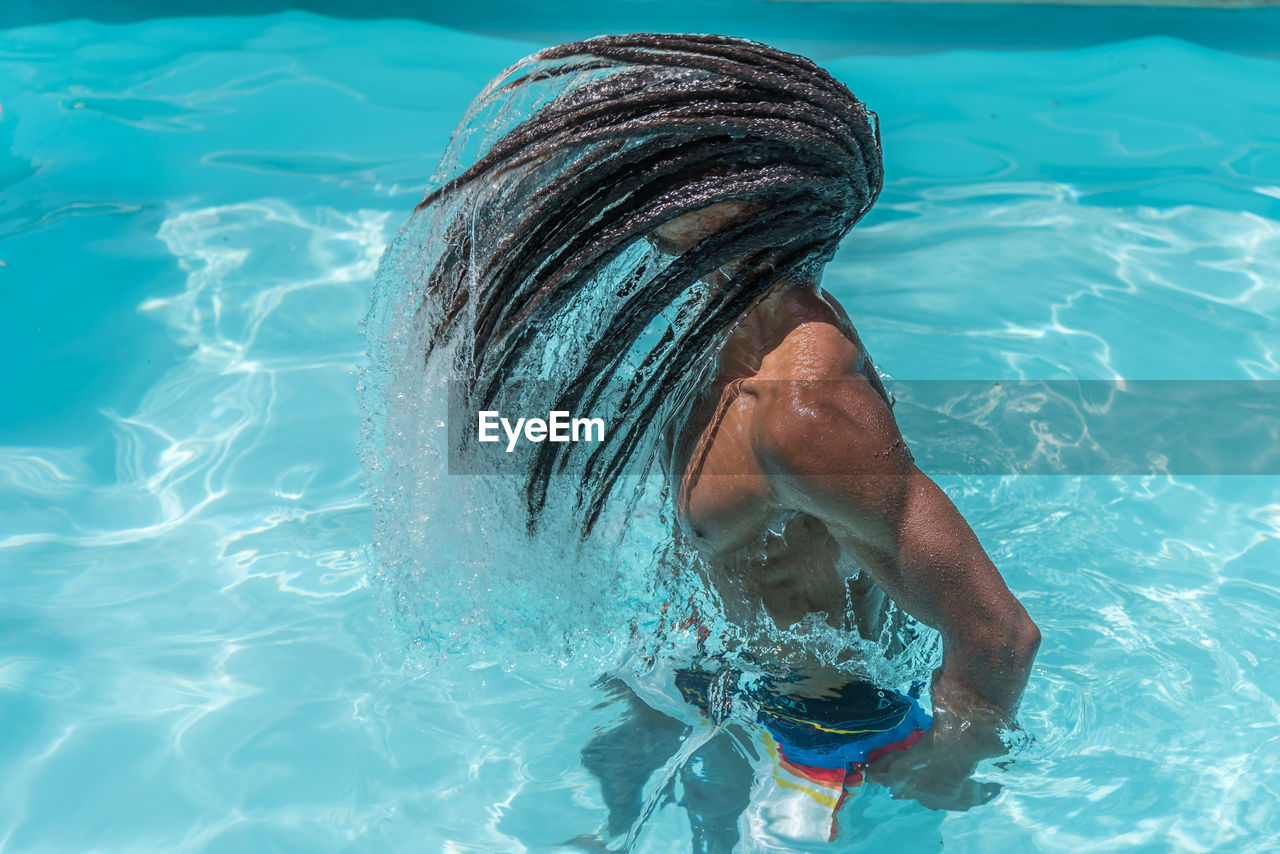 Young black man with dreadlocks inside a pool moving his wet hair in a trail of water