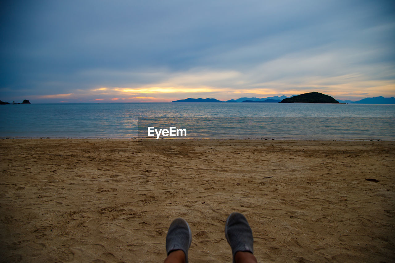 sea, water, land, sky, beach, ocean, horizon, sunset, low section, sunlight, cloud, sand, human leg, body of water, nature, coast, scenics - nature, beauty in nature, shore, personal perspective, wave, shoe, one person, reflection, evening, dusk, relaxation, blue, tranquility, human foot, leisure activity, horizon over water, trip, vacation, holiday, lifestyles, tranquil scene, travel destinations, outdoors, limb, human limb, travel, rock, sun, coastline, idyllic, mountain, tourism, adult, standing