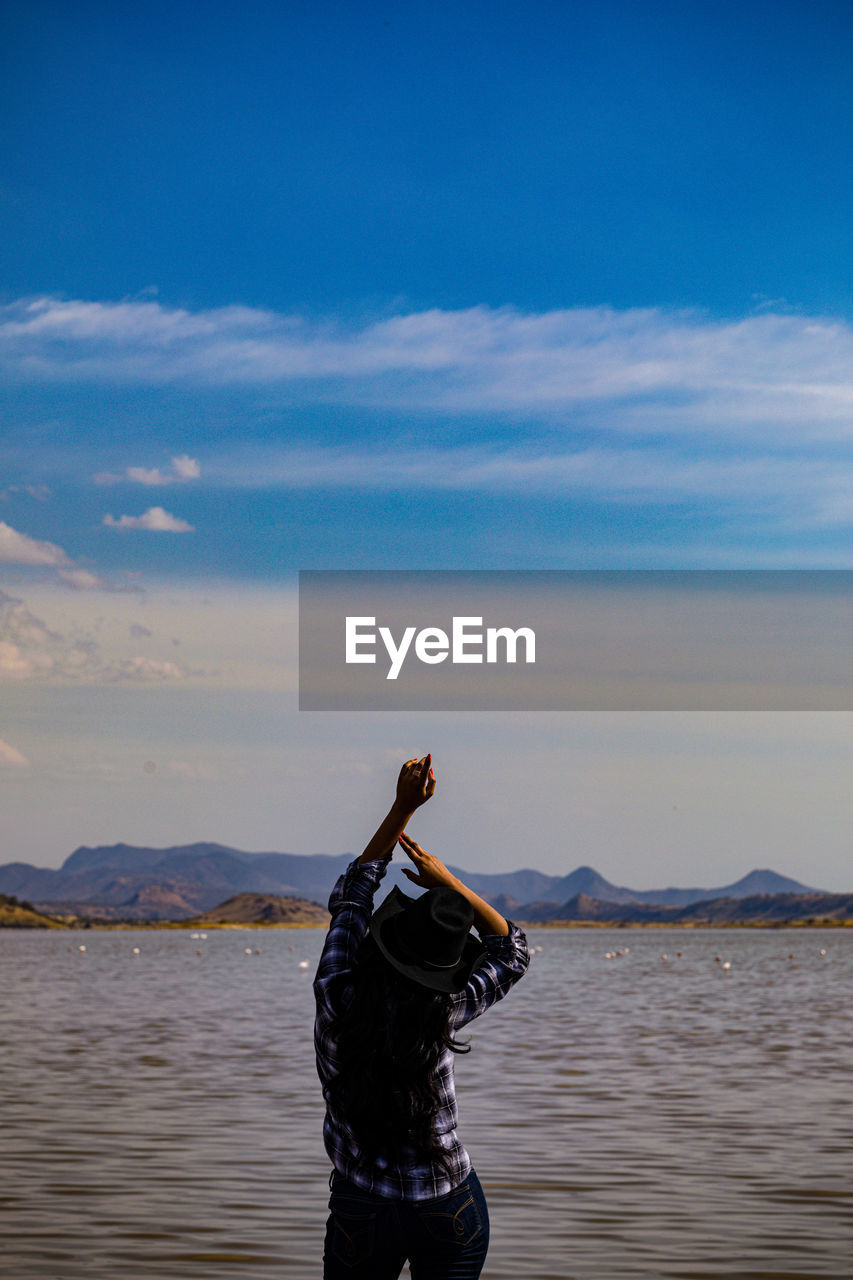 water, sky, sea, one person, nature, mountain, beauty in nature, cloud, blue, ocean, arm, scenics - nature, beach, adult, standing, leisure activity, reflection, rear view, land, shore, arms raised, lifestyles, outdoors, three quarter length, men, holiday, limb, day, tranquility, vacation, trip, horizon, coast, environment, wave, tranquil scene, human limb, sports, mountain range, travel, full length