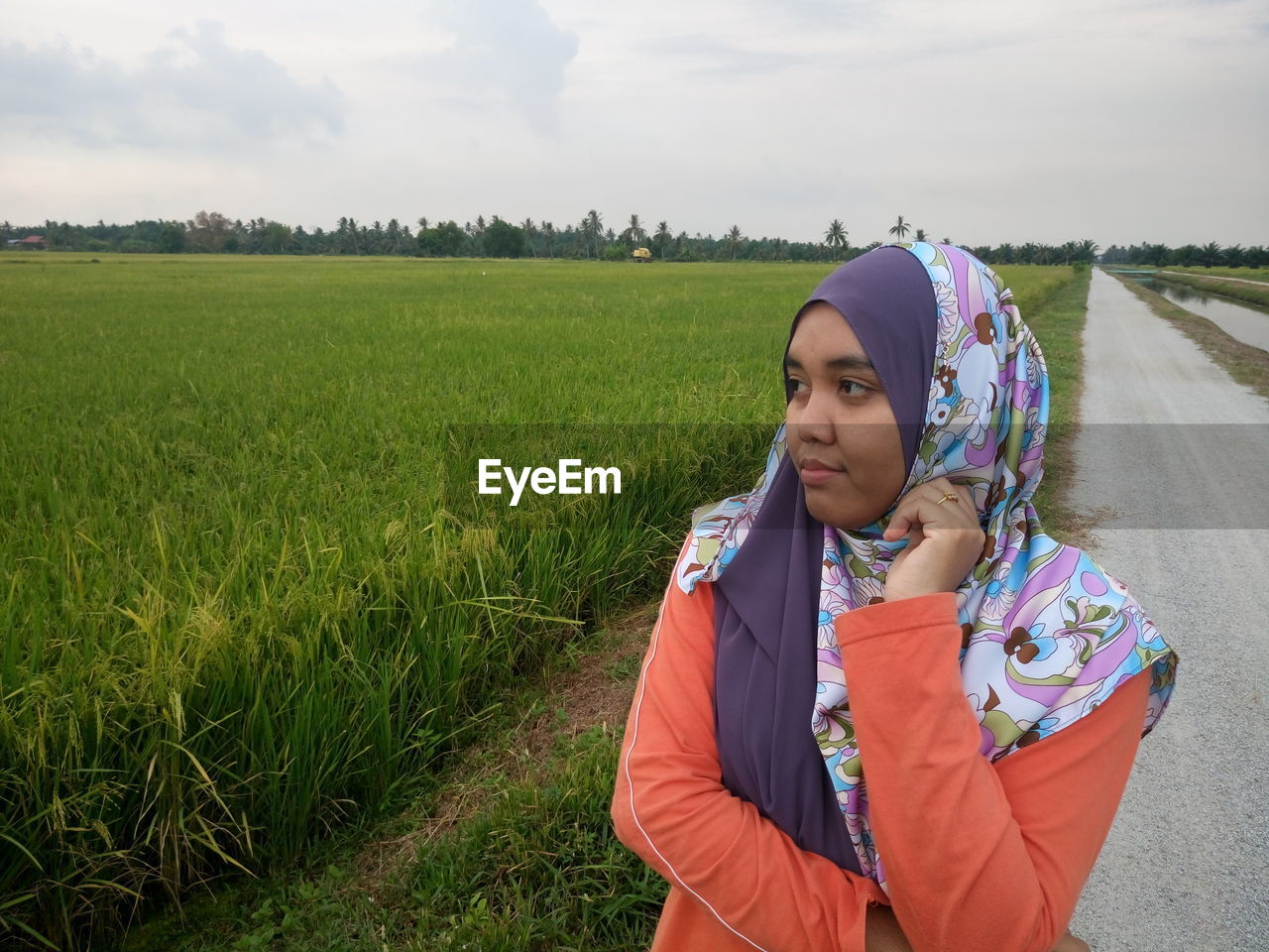 Woman in hijab looking away while standing on country road by rice paddy field