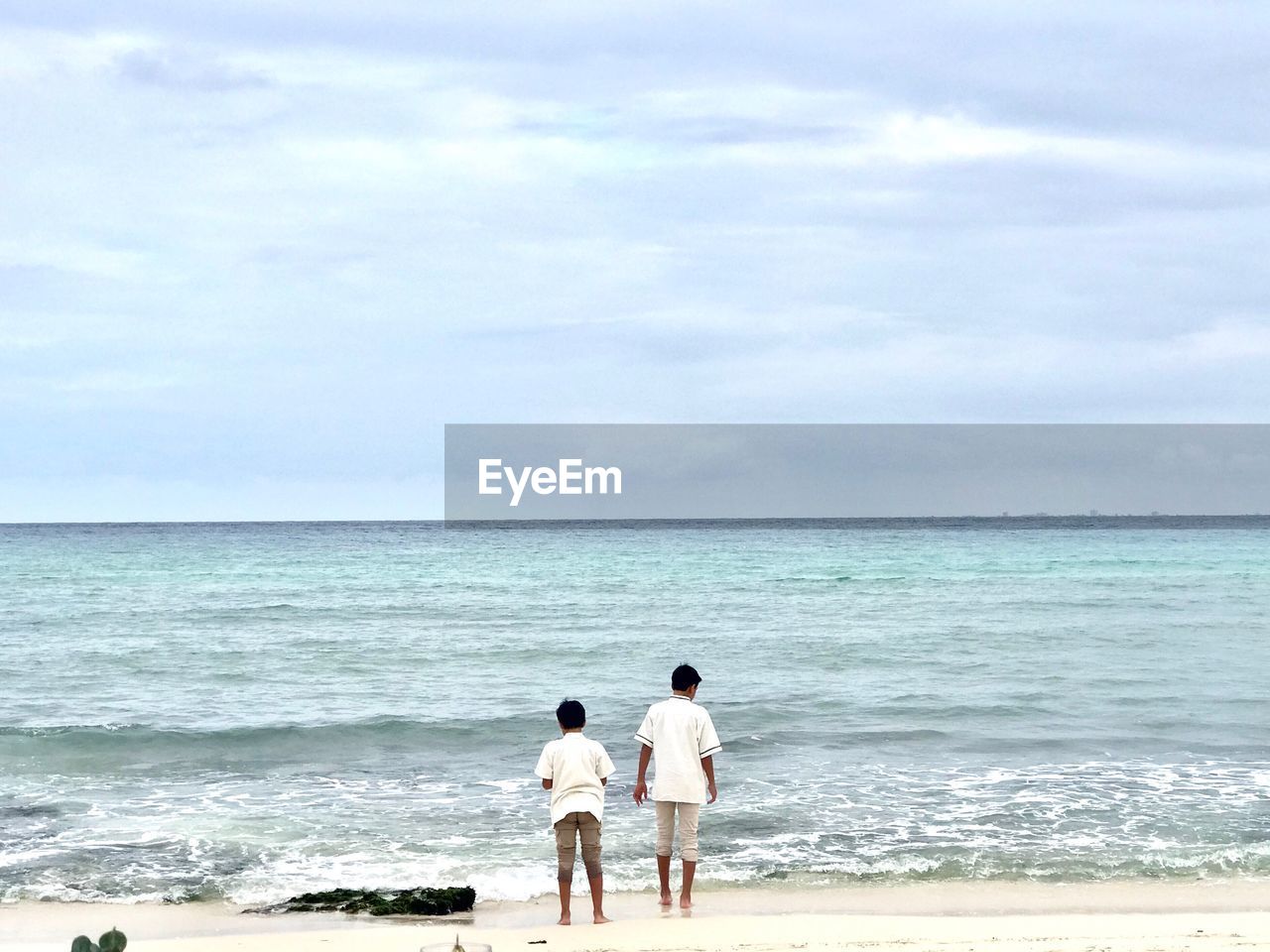 Rear view of male friends standing at beach against cloudy sky