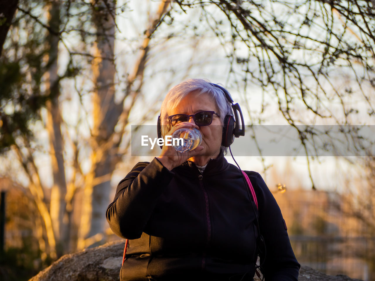 Senior woman drinking water from bottle in park during sunset