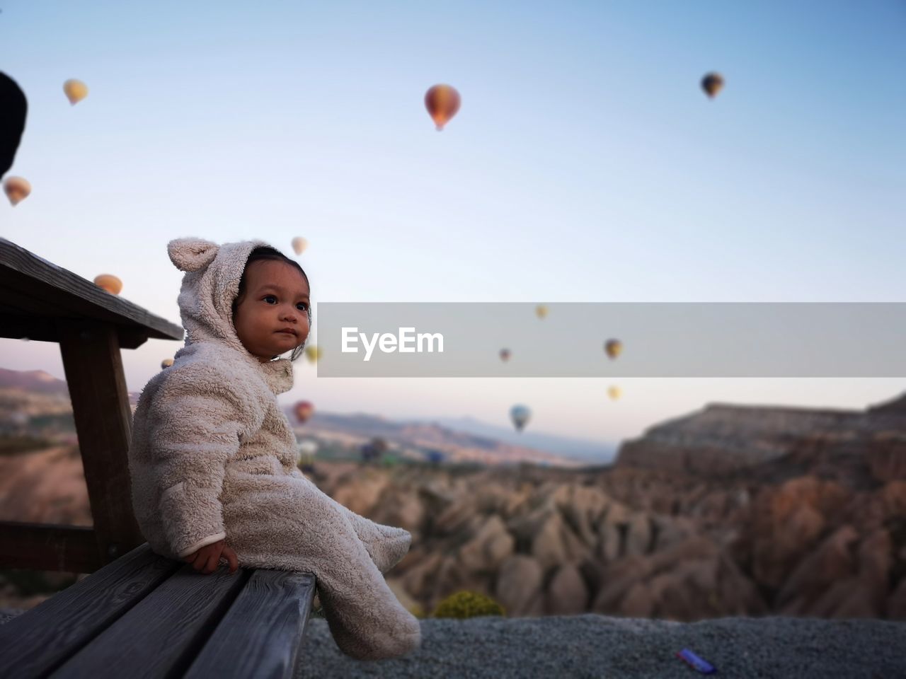 Baby girl sitting on bench against hot air balloons flying over mountains