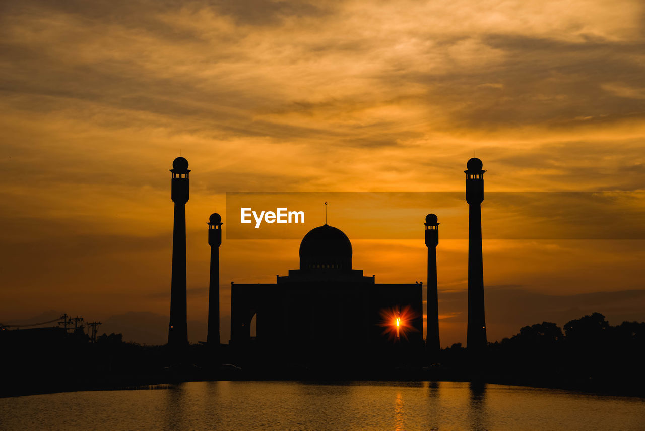 Silhouette mosque by lake against orange sky
