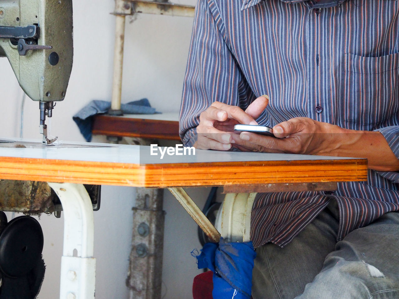 Midsection of man using mobile phone while sitting by sewing machine