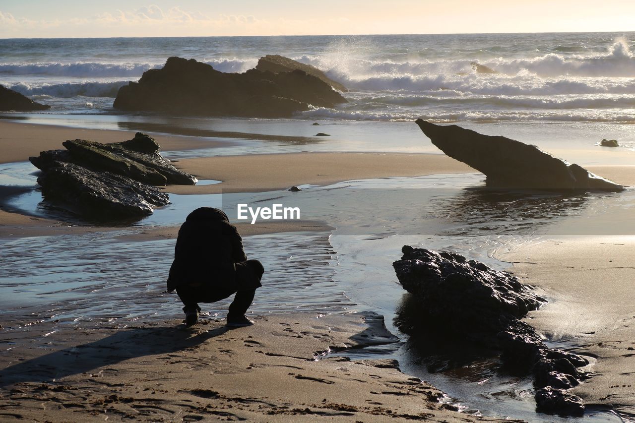 Rear view of man crouching at beach during sunset