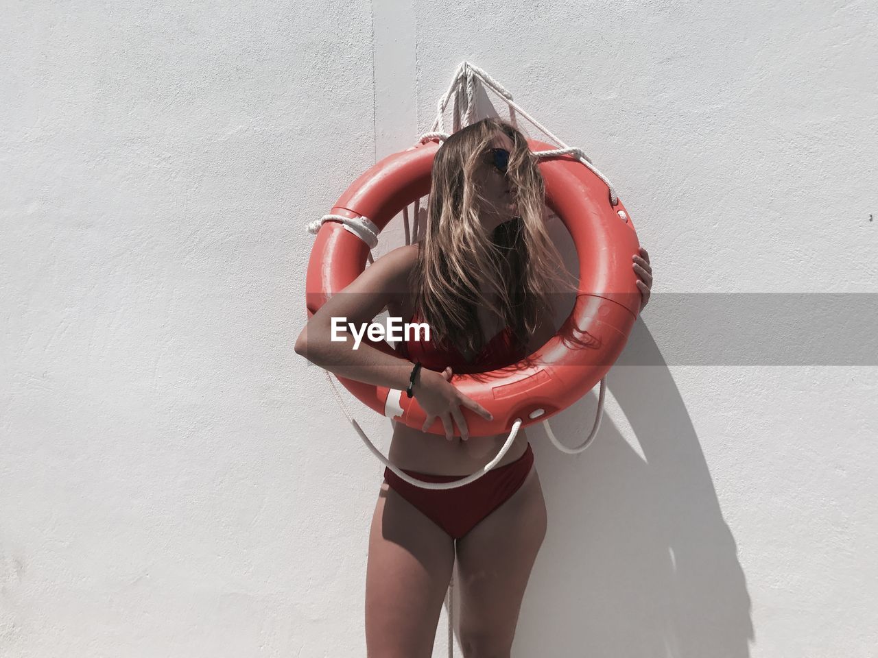 Carefree young woman in bikini covered with inflatable ring standing against wall