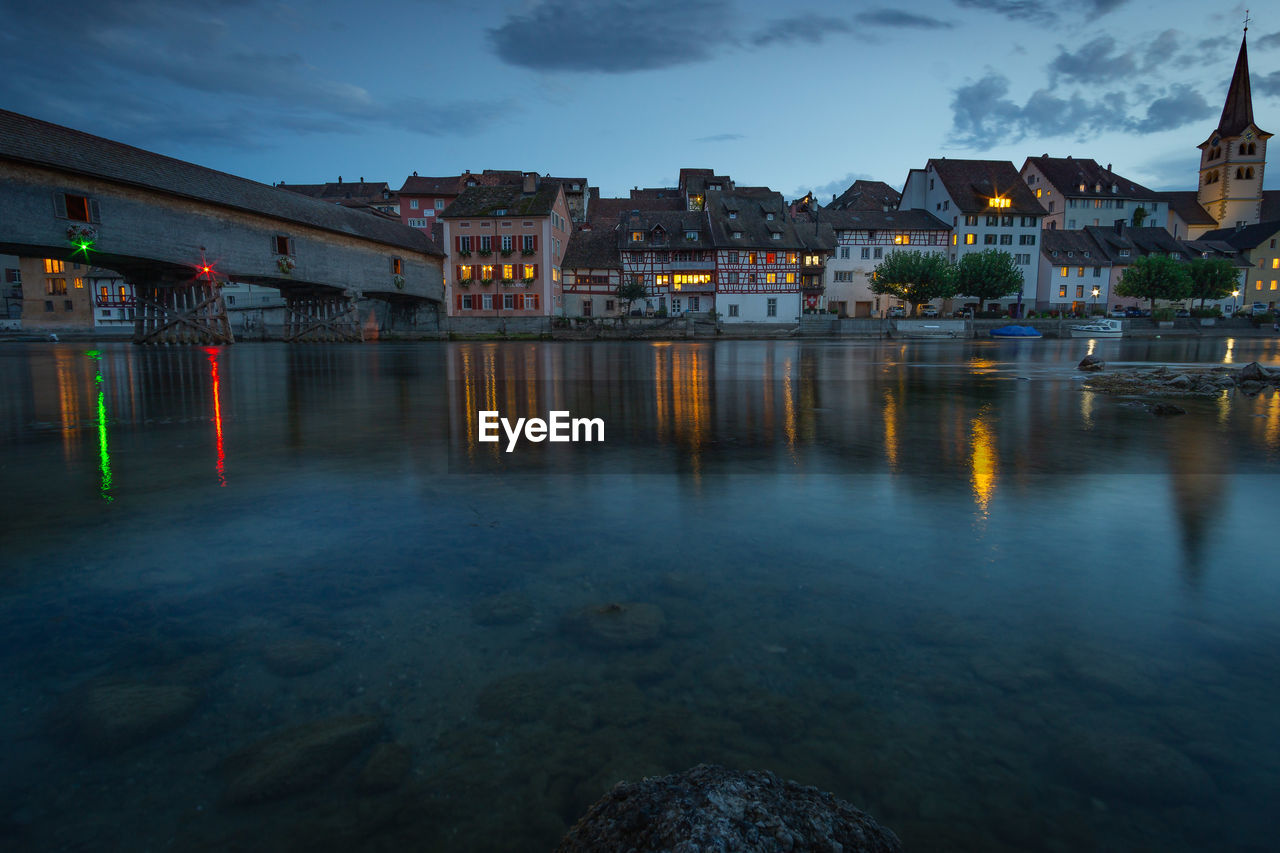 Illuminated buildings by river against sky at dusk