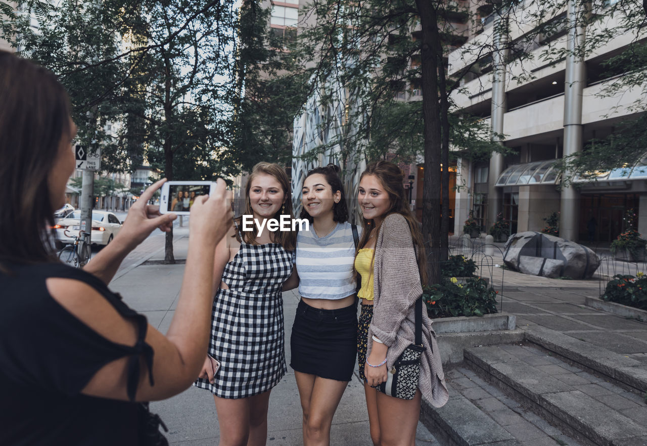 Side view of woman photographing friends with smart phone on sidewalk