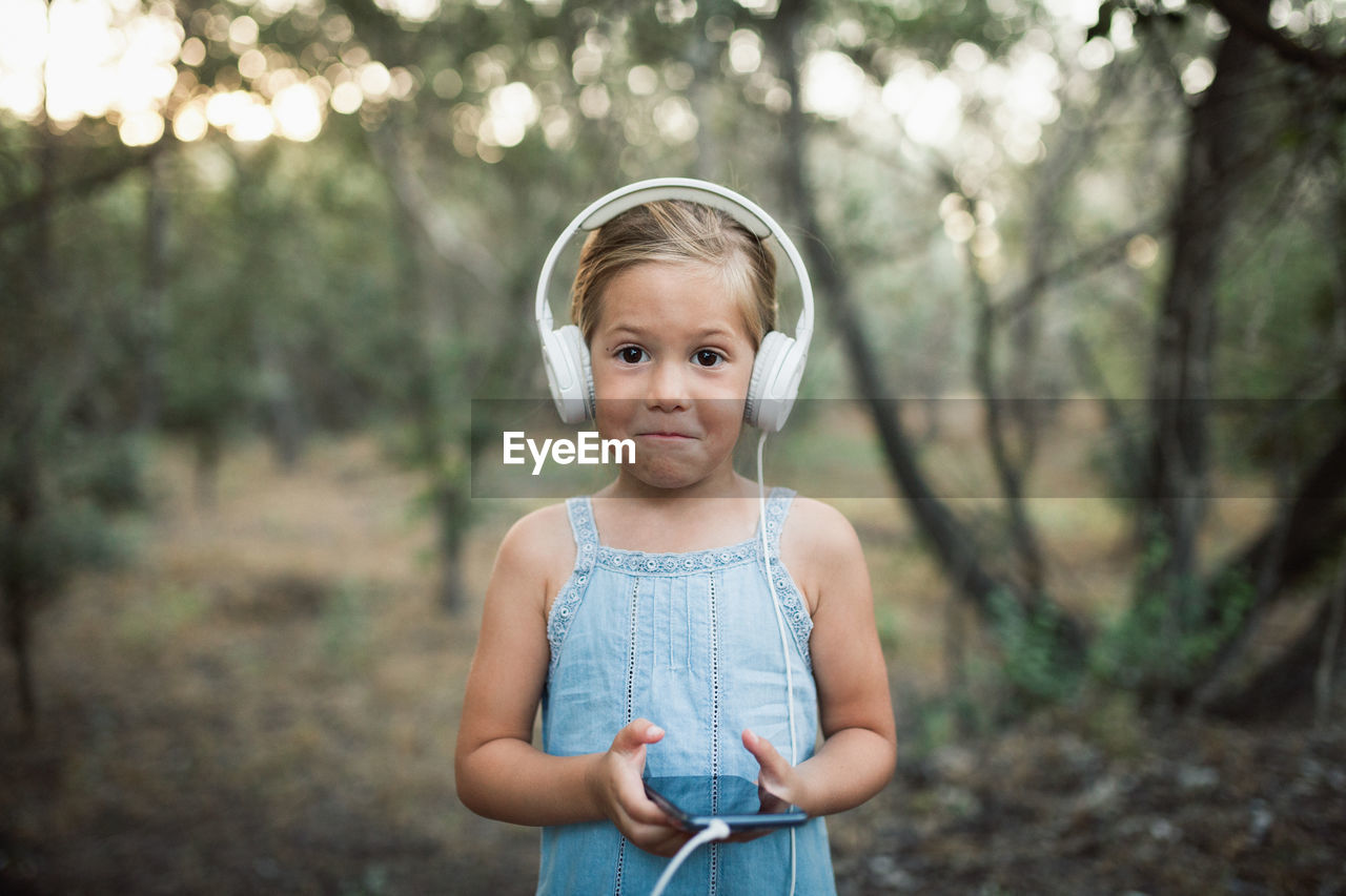 Portrait of cute girl listening to music standing against trees