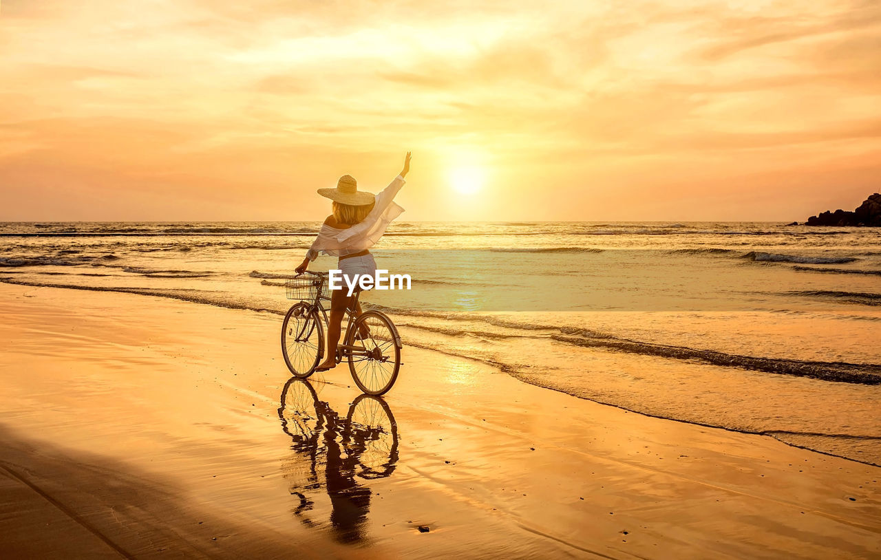Woman traveler with bicycle walking sea coastline, looking on sunset sky, sunny summer day on beach.