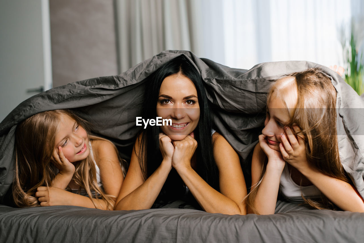 Mom and two daughters are lying on the bed under a blanket. the brunette mother looks at the camera